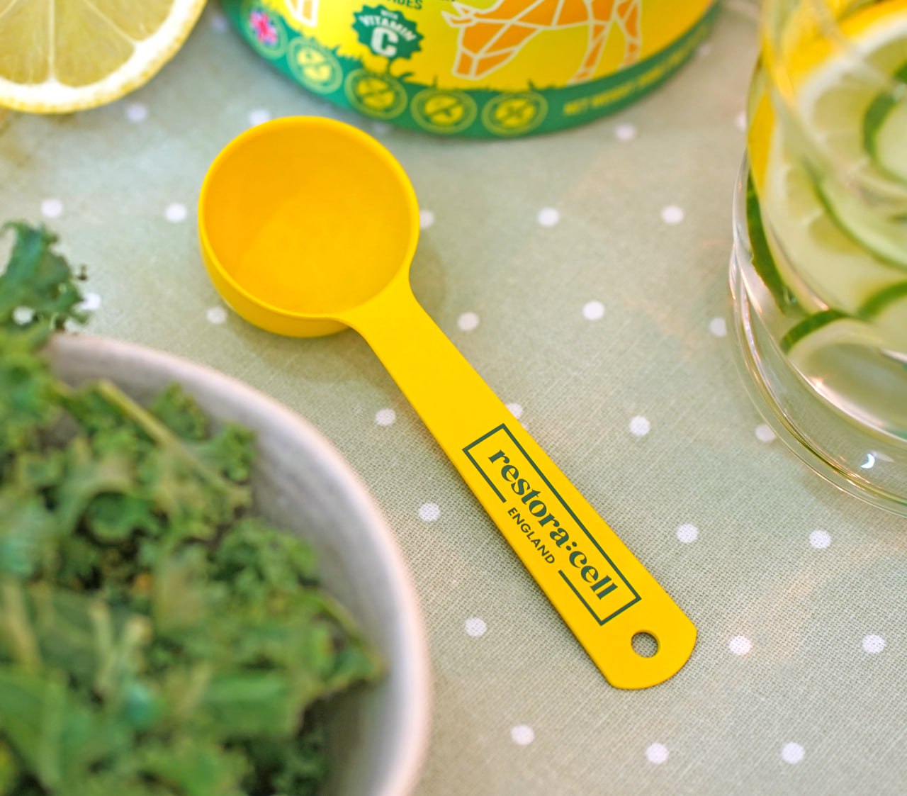 Bright yellow Restora:cell supplement scoop on a green polka dot tablecloth, surrounded by fresh kale, cucumber water, and a lemon slice – health and wellness lifestyle concept.
