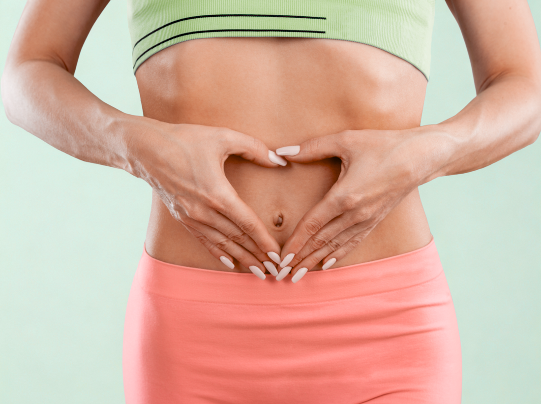 A woman wearing a green sports top and pink leggings forms a heart shape with her hands over her stomach, symbolizing gut health and wellness.