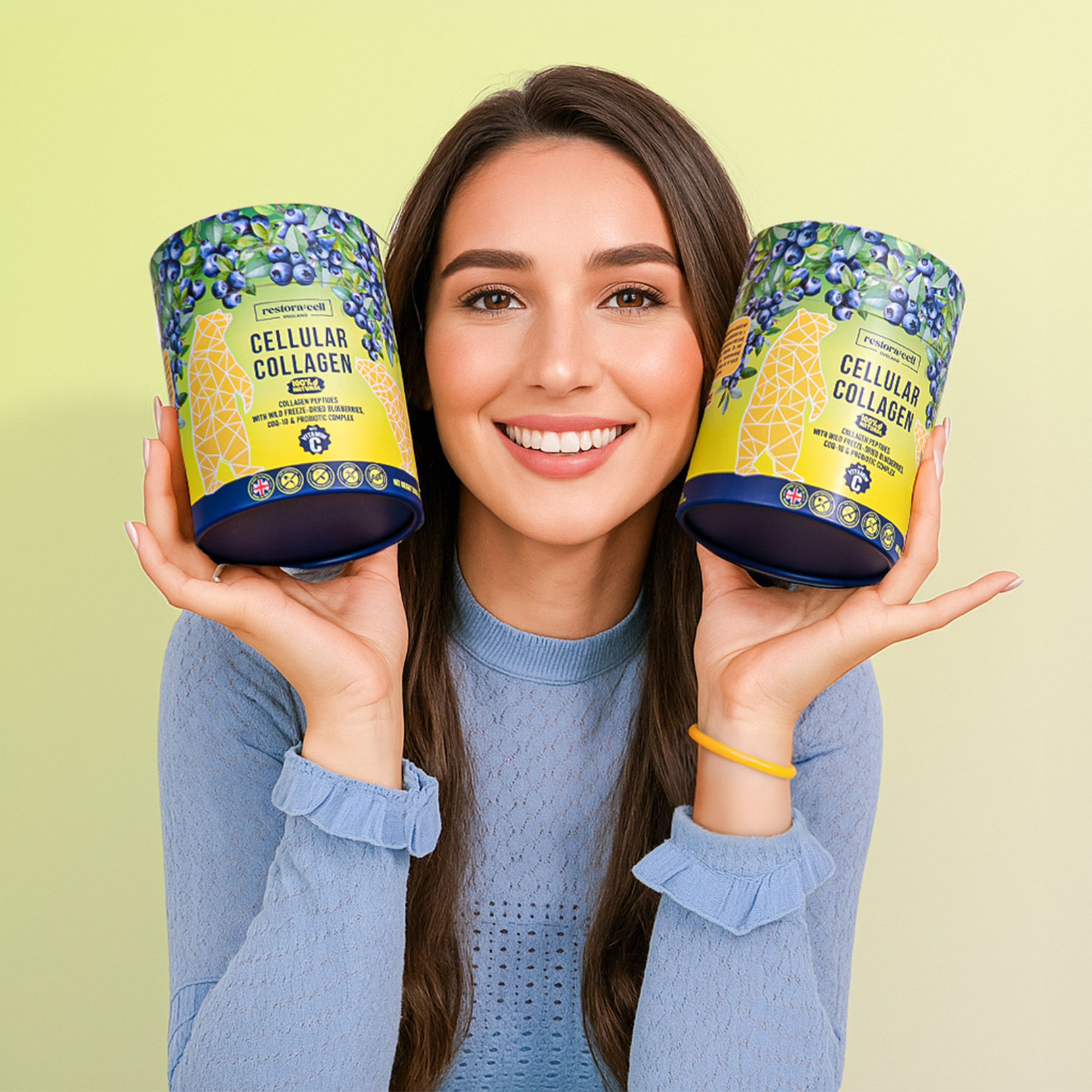A smiling woman with long brown hair holds two colorful containers of Restora:Cell Cellular Collagen Powder beside her face. She wears a light blue sweater and poses against a soft yellow-green background.
