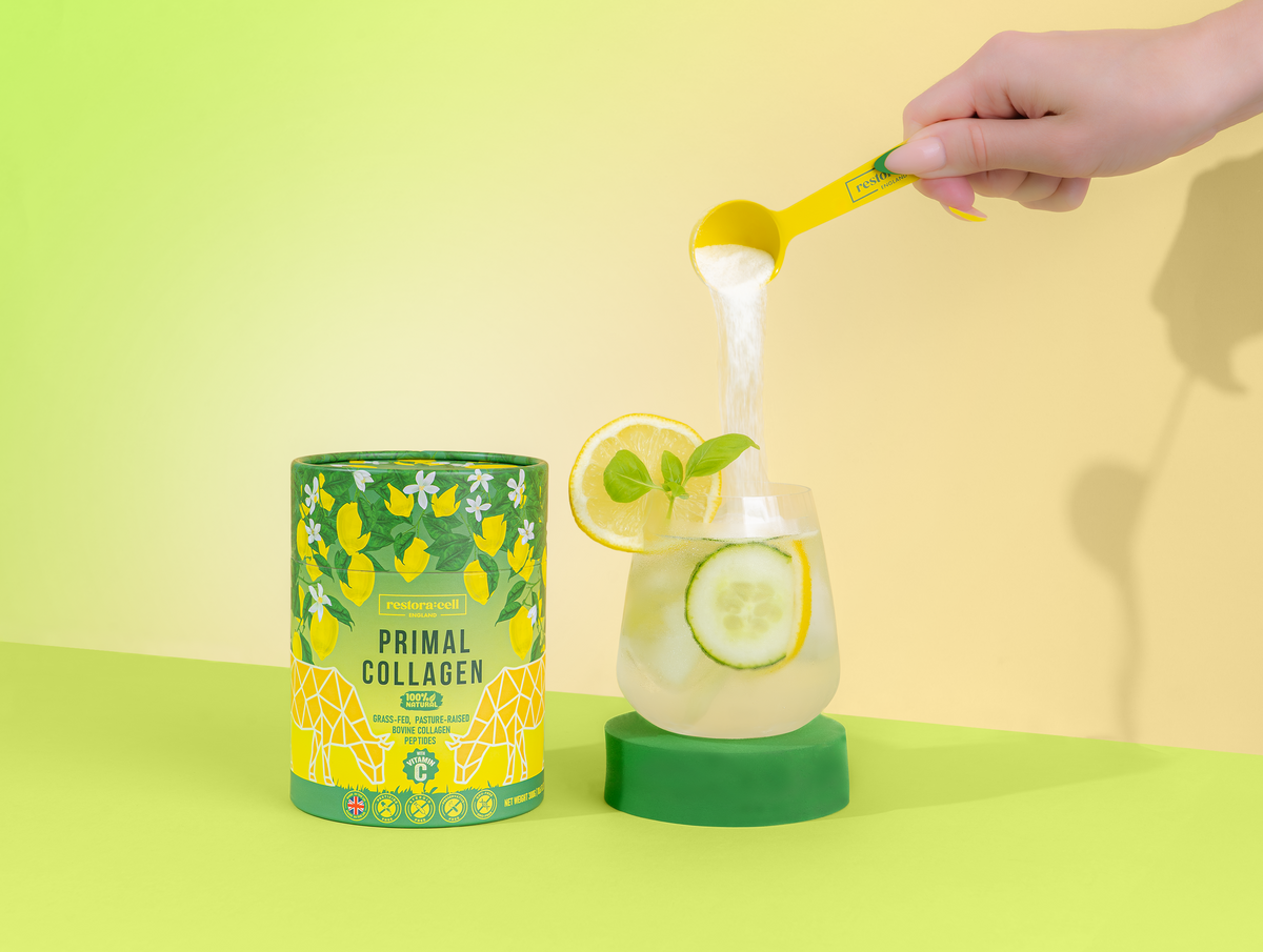 restora:cell Primal Collagen beside a glass with lemon, cucumber and mint, as powder is poured in from a yellow scoop.